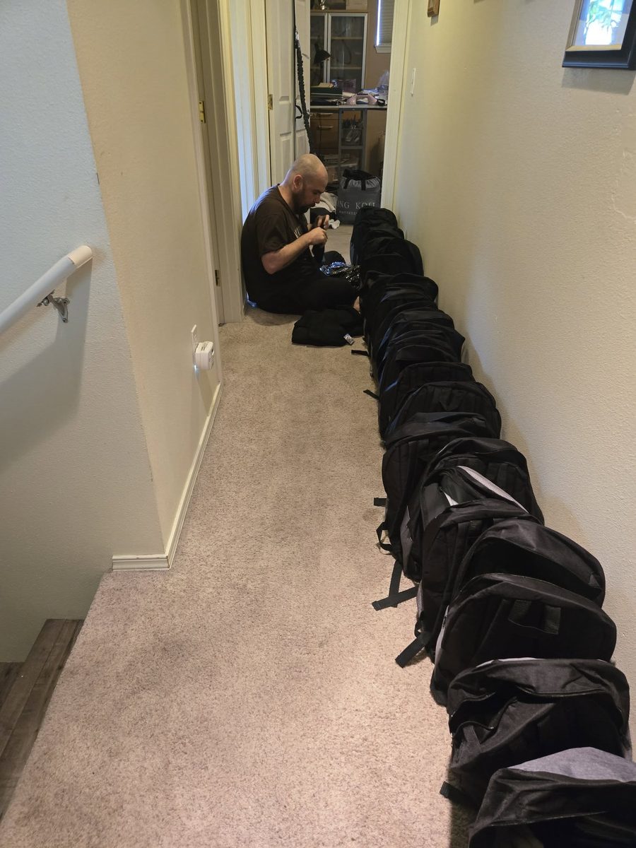 Michael, in a brown t-shirt, sits cross-legged on the carpet at the far end of a hallway, filling about twenty black backpacks lined up in a long row that leads toward the camera. Loose supplies and a foil emergency blanket rest on the floor beside him. The photo looks down the hallway toward him from the near end; a white handrail runs along the left wall and a framed picture hangs by the doorway behind him. The overall feel is quiet and focused &mdash; one person doing a large job by hand.